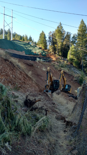 Excavators on hillside near forest and power lines.